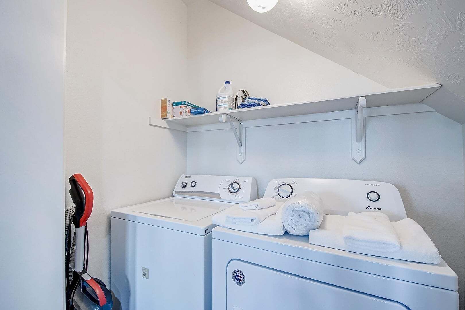 A laundry area with two washing machines, a shelf with laundry supplies, and a vacuum cleaner.