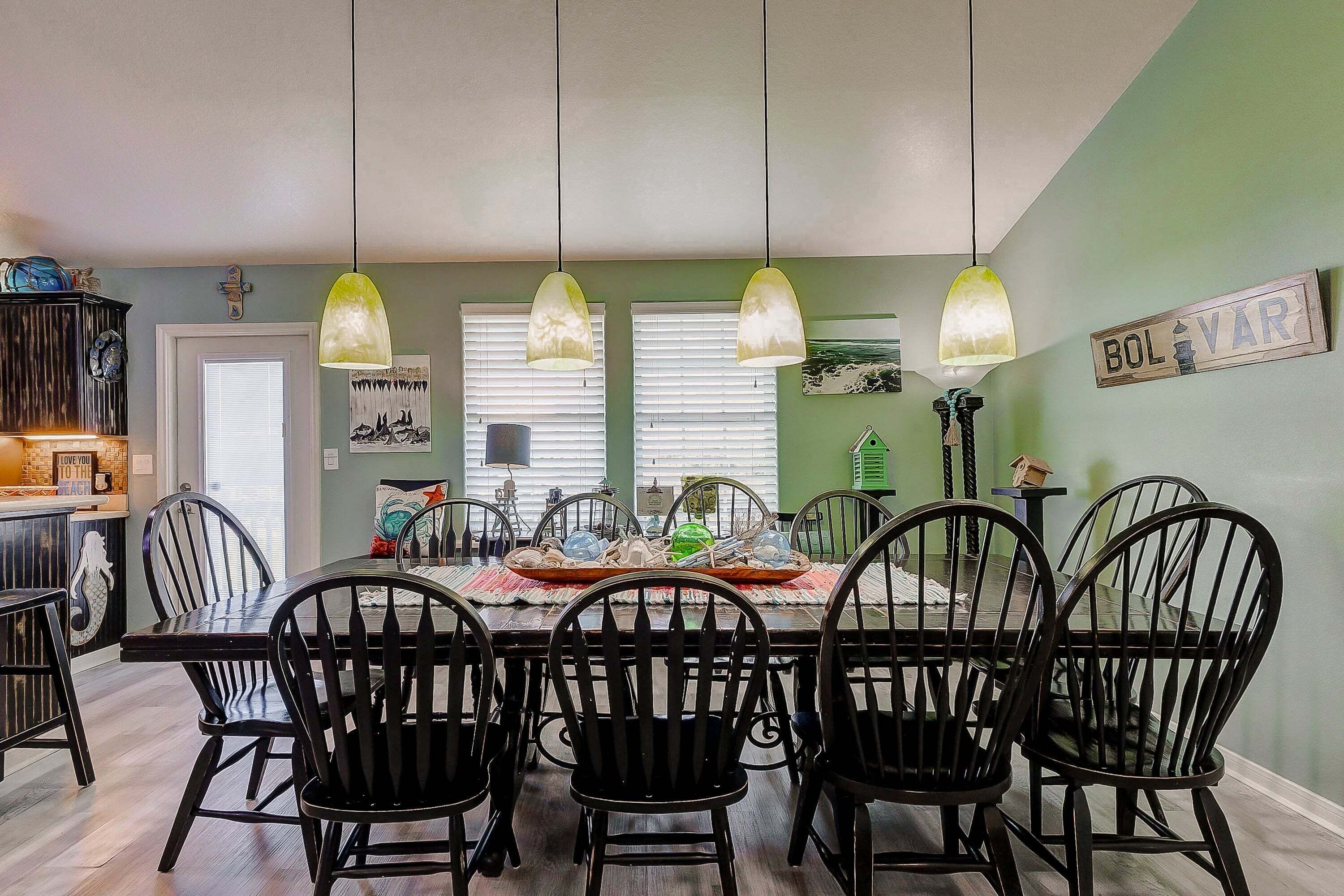 A dining area with a table set for six, black chairs, hanging pendant lights, and a green wall with a sign that reads 'BOLVAR'.
