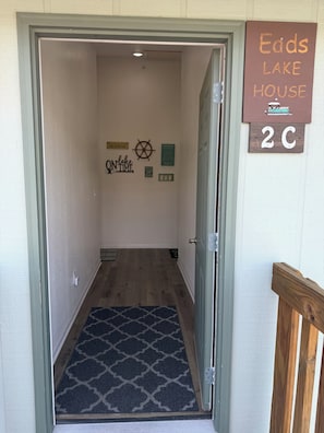 Ein Flur mit Fußmatte, einem Schild mit der Aufschrift 'Eads Lake House 2C' und Wanddekorationen, darunter ein Schiffsschwengel und ein Schild, das 'Am See Zeit' besagt.