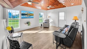 A spacious room with a wooden ceiling, a large TV, a sofa, a dining table, and a view of a lake. - Over The Rainbow 3 Bedroom Home (Port Bolivar)