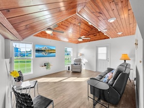 A spacious room with a wooden ceiling, a large TV, a sofa, a dining table, and a view of a lake. - Over The Rainbow 3 Bedroom Home (Port Bolivar)