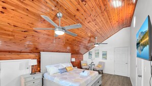 A bedroom with a sloped ceiling, wooden paneling, a bed with a patterned bedspread, two bedside tables, a nightstand, a ceiling fan, and a large window with a mountain view. - Over The Rainbow 3 Bedroom Home (Port Bolivar)