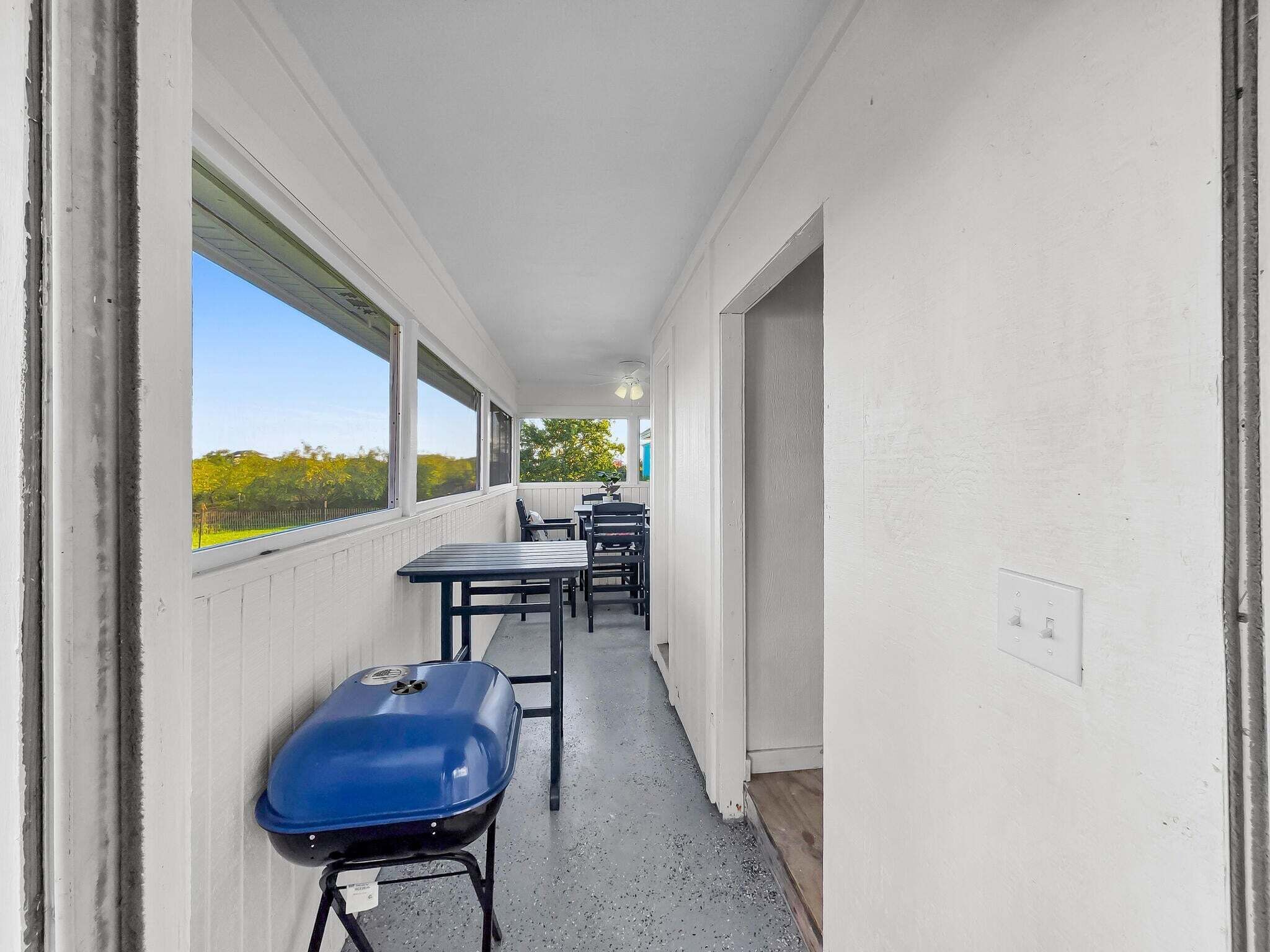 A narrow corridor with a covered area featuring tables and chairs, a blue grill, and a view of greenery outside.