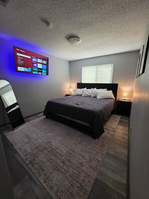 A modern bedroom with a bed, bedside lamps, a TV mounted on the wall, and a window with blinds. - Modern Home with Backyard pool + Mini Golf! (San Antonio)