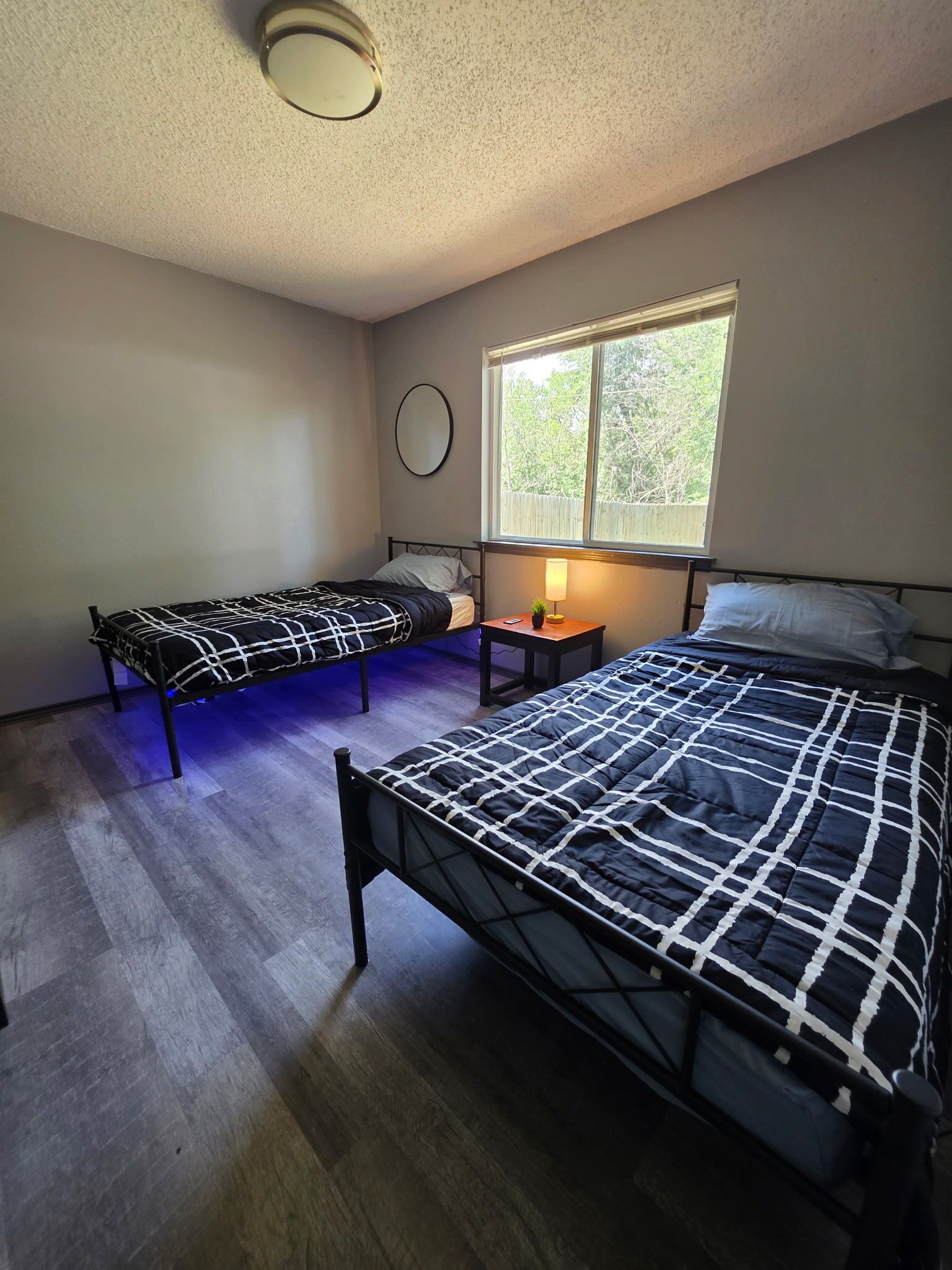 Une chambre avec deux lits simples, une table de chevet avec une lampe, un miroir rond et une fenêtre donnant sur des arbres.
