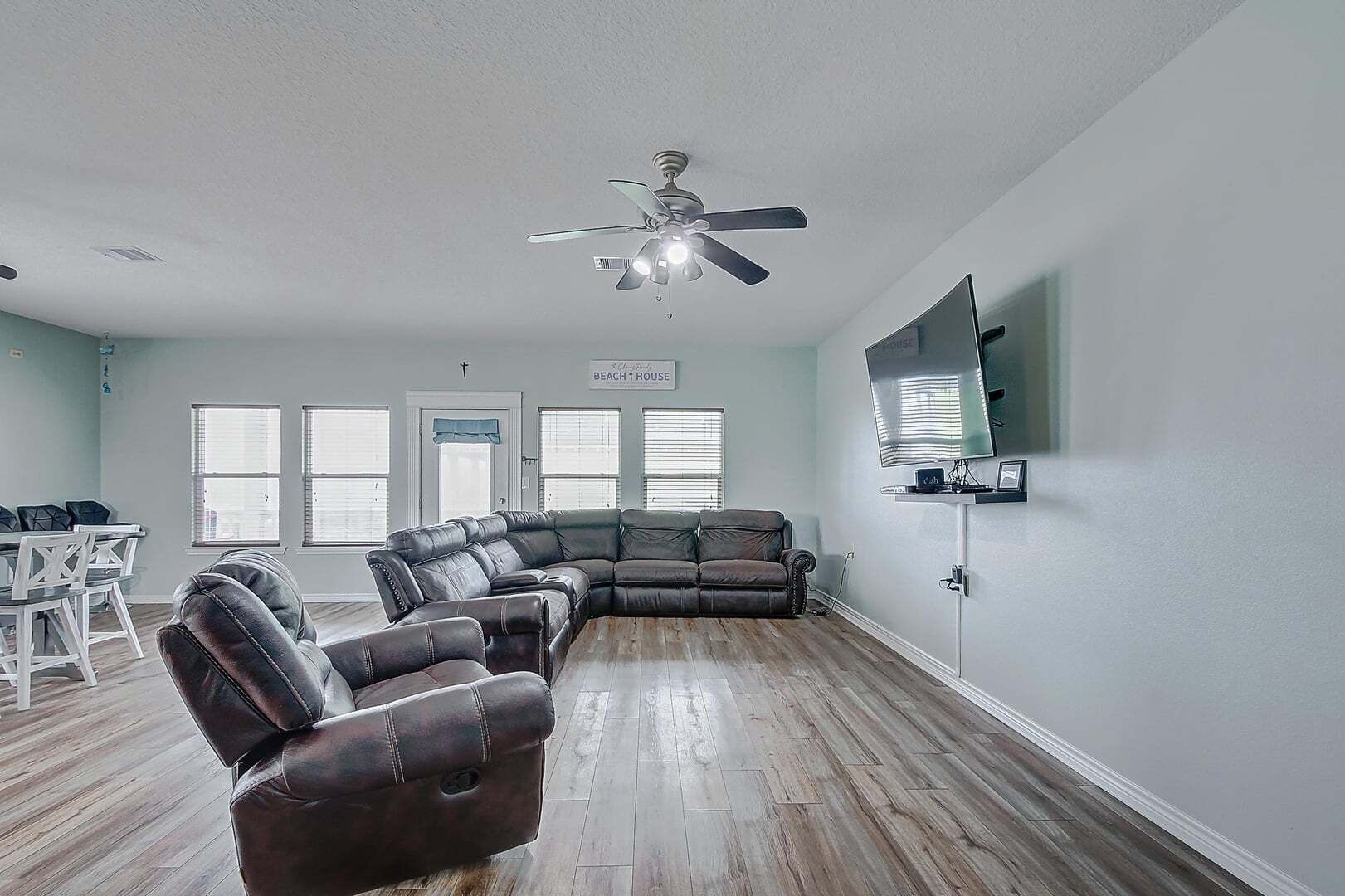 A living room with a sectional sofa, a ceiling fan, a flat-screen TV, and a dining area with a white table and chairs.