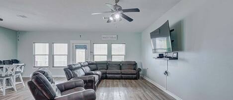 A living room with a sectional sofa, a ceiling fan, a flat-screen TV, and a dining area with a white table and chairs.