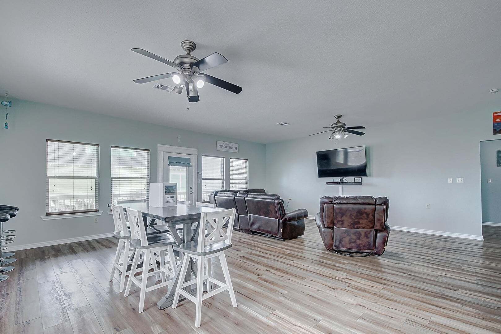 Un salón amplio con comedor, dos ventiladores de techo, un televisor de pantalla plana y un letrero que dice 'BEACH HOUSE'.