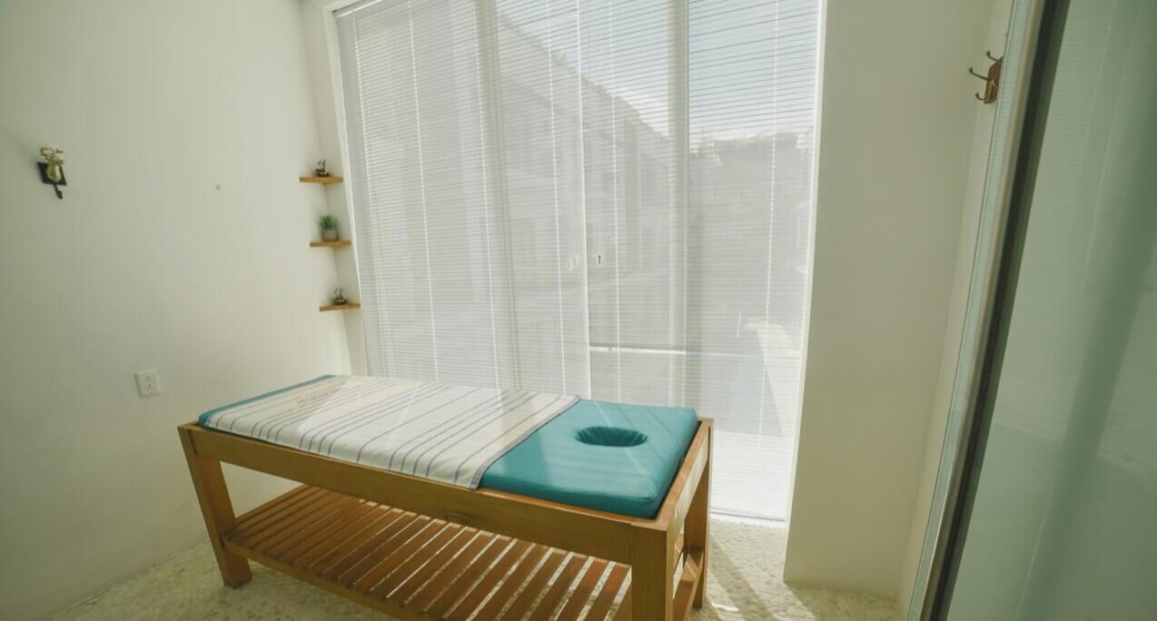 A small room with a single bed, a window with blinds, a wooden floor, and a small shelf with items.