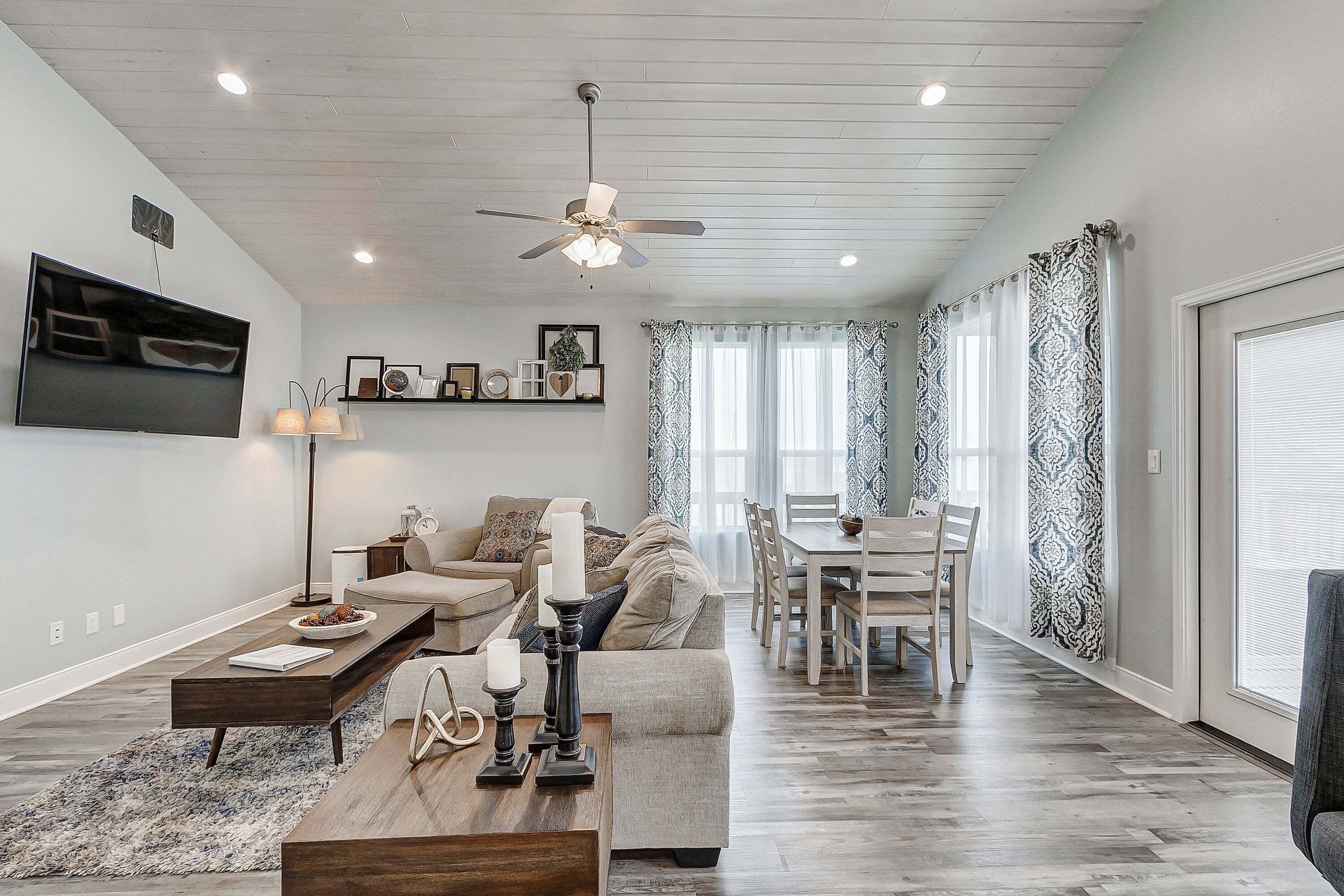 A modern living room with a flat-screen TV, a ceiling fan, a dining area with a table and chairs, and patterned curtains.