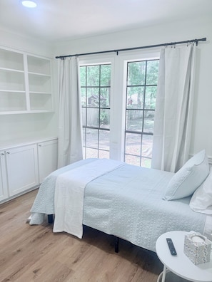Une chambre à coucher avec un lit, une fenêtre avec des rideaux, une table de chevet blanche et une télécommande.