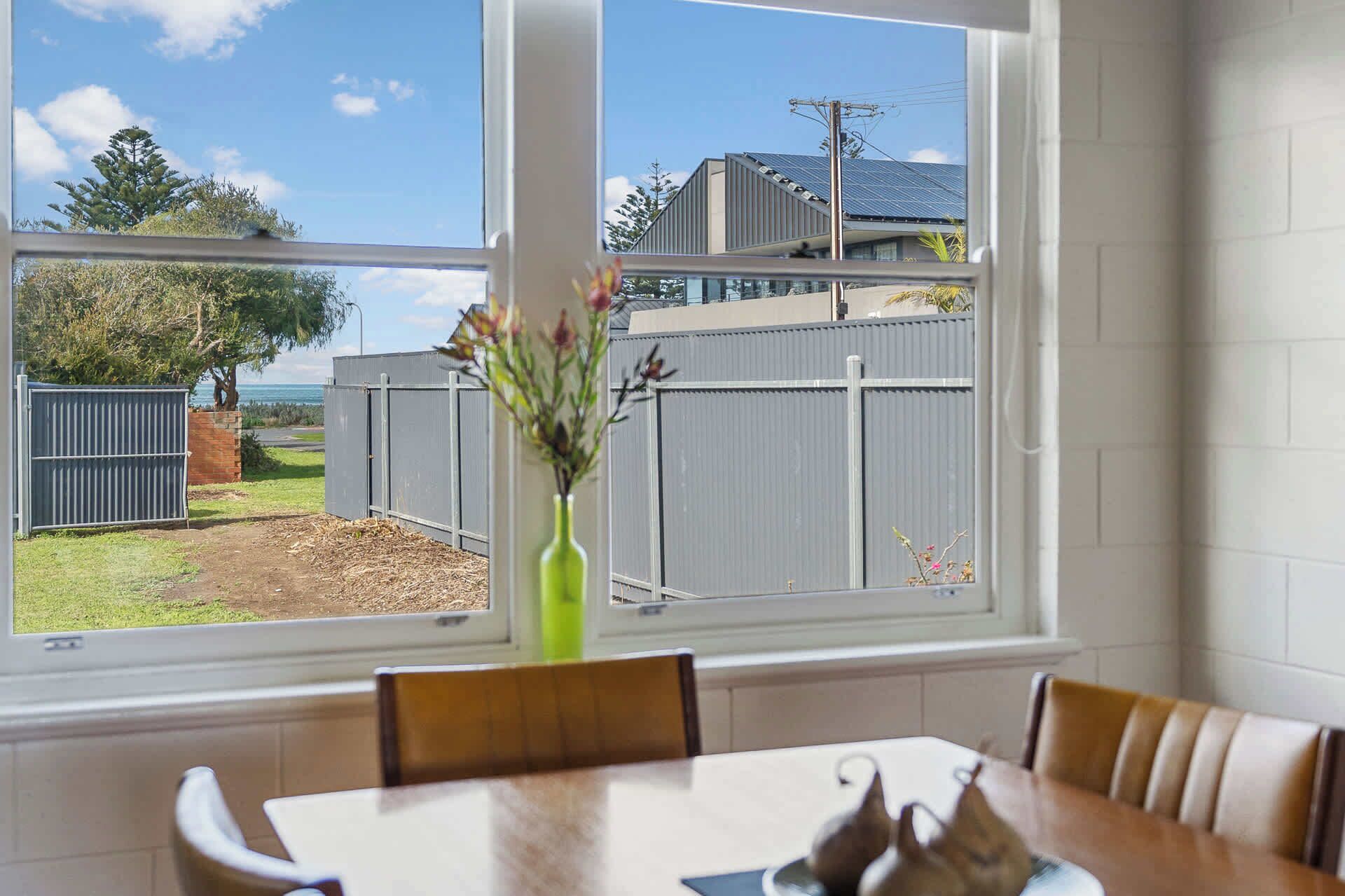 A dining area with a table and chairs, a window with a view of a fenced backyard and a building with solar panels.