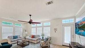 A spacious living room with a ceiling fan, large windows, a television, and a fireplace. - Mermaid Mansion 3 Bedroom Home (Port Bolivar)