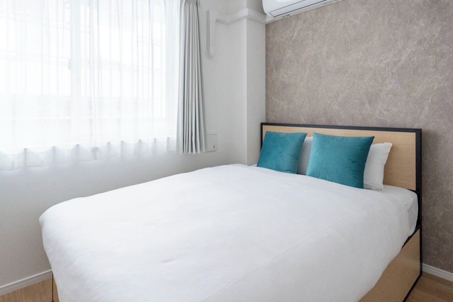 Cama bem arrumada com roupa de cama branca e dois travesseiros azul-esverdeados, janela com cortinas leves e parede texturizada atrás da cama.