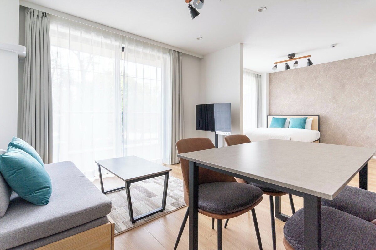 A modern hotel room with a sofa, a dining table, chairs, a TV, and a bed with a headboard.