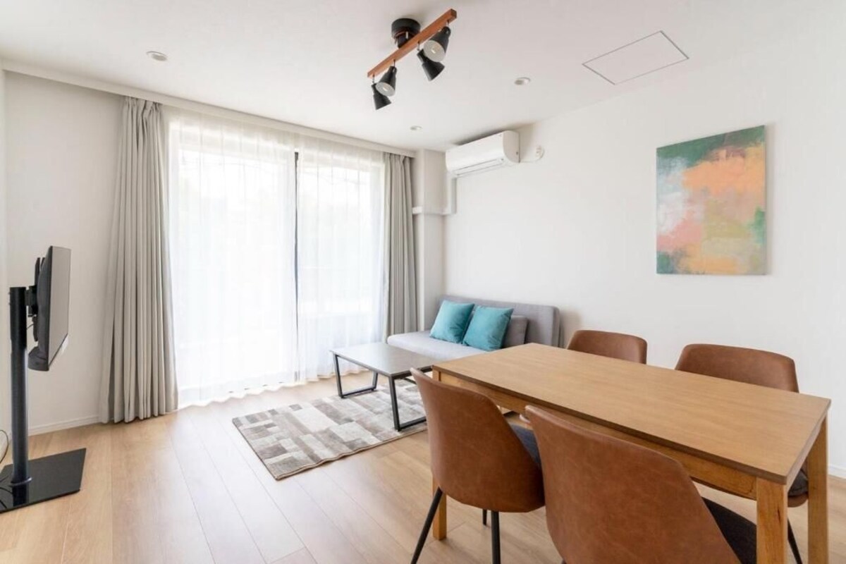 A modern living room with a wooden dining table, brown chairs, a grey sofa with blue cushions, a flat-screen TV, and a large window with curtains.