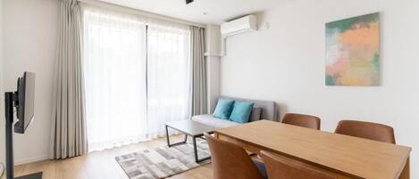 A modern living room with a wooden dining table, brown chairs, a grey sofa with blue cushions, a flat-screen TV, and a large window with curtains.