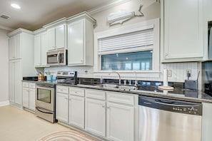 A kitchen with white cabinets, a stainless steel refrigerator, a microwave, a dishwasher, and a window with blinds. - Blessings By The Sea 3 Bedroom Home (Port Bolivar)