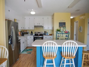 A kitchen with white cabinets, a stainless steel refrigerator, a microwave, and a stove. There are two white bar stools at the island. - Sand in my boots - 2 Bedroom home in GULF SHORES with private pool and bikes (Gulf Shores)
