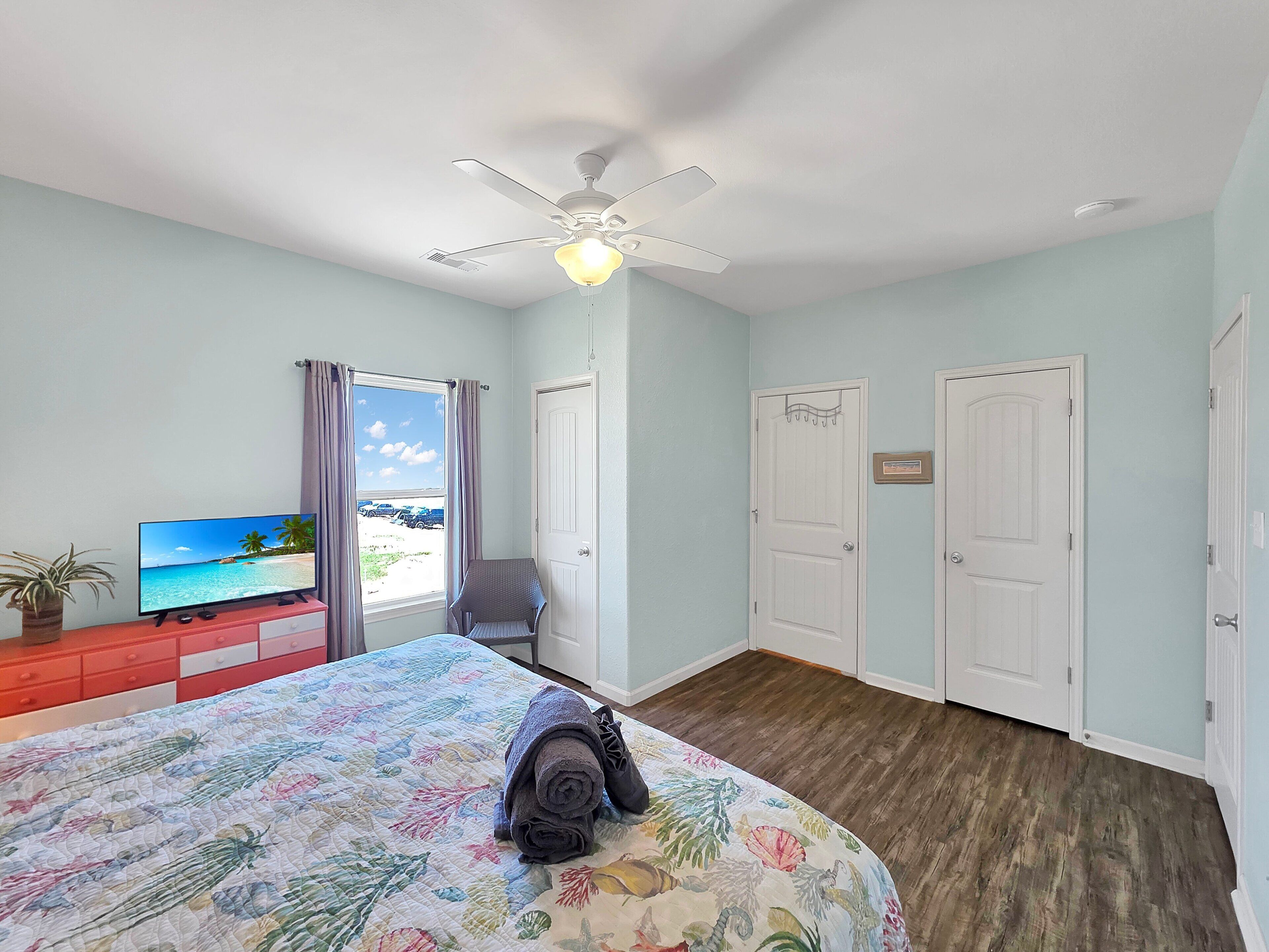 A hotel room with a bed, a television, a ceiling fan, and a view of the beach through the window.