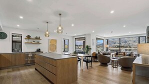 A modern kitchen with wooden cabinets, a central island, pendant lighting, and large windows offering a view of mountains. - Lakeside Bliss Luxury 3 Bedroom Apartment (Wānaka)