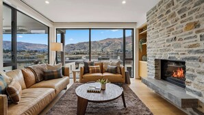 A modern living room with leather sofas, a coffee table, a fireplace, and a large window with a mountain view. - Lakeside Bliss Luxury 3 Bedroom Apartment (Wānaka)