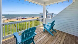 A covered porch with two chairs, a view of the beach, and a building in the background. - Blue Anchor 5 Bedroom Home (Port Bolivar)