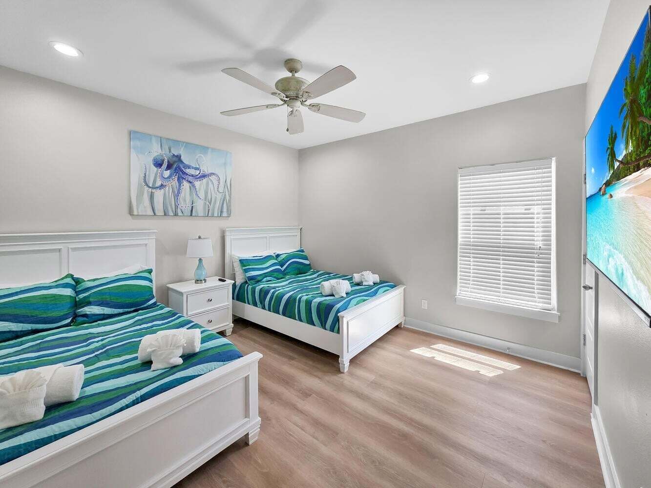 A bedroom with two beds, a ceiling fan, a painting, a window with blinds, and a TV.