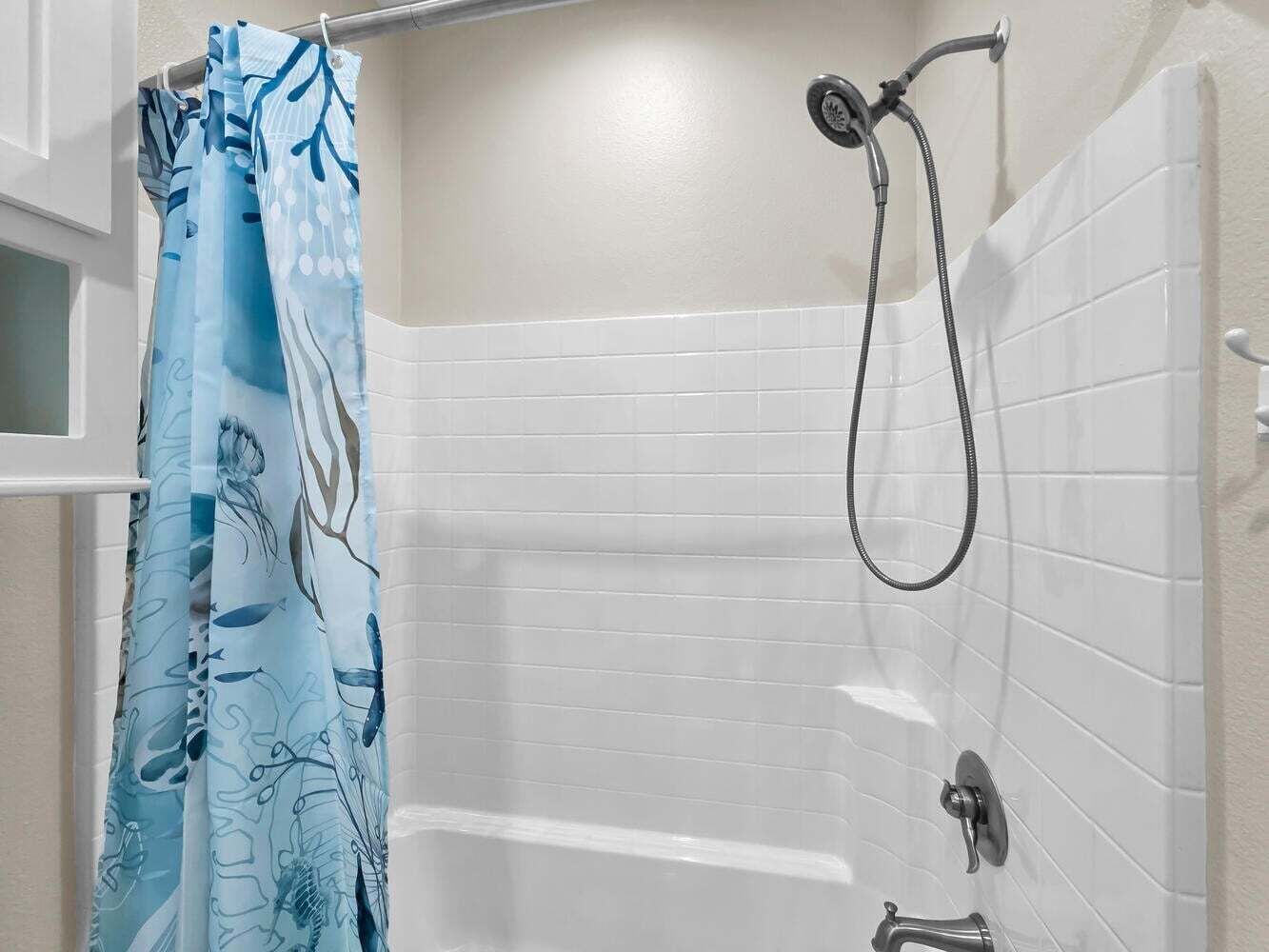 A bathroom with white tiled walls, a shower with a handheld showerhead, and a blue shower curtain with a floral pattern.