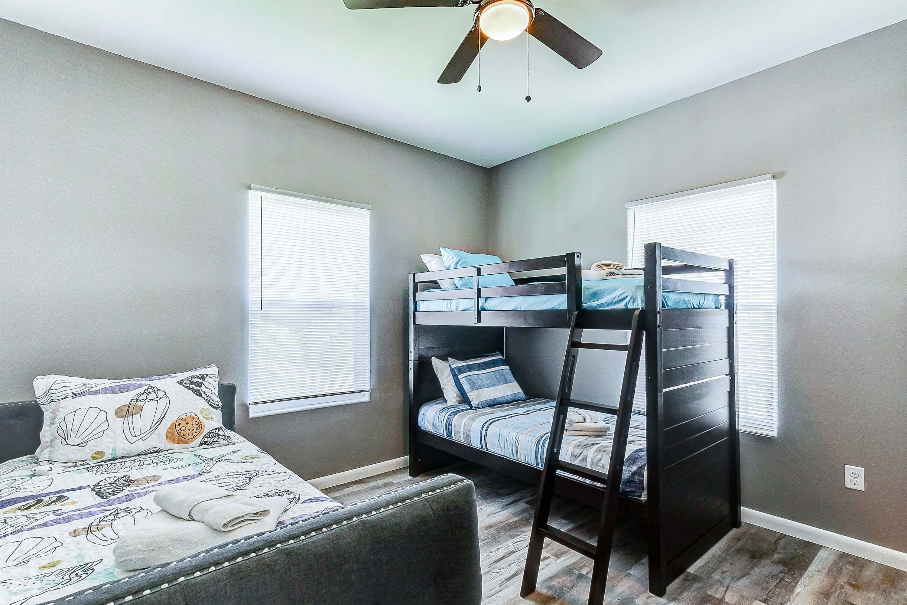 Quarto com cama de solteiro, uma cama individual com roupa de cama estampada, uma cadeira e um ventilador de teto.