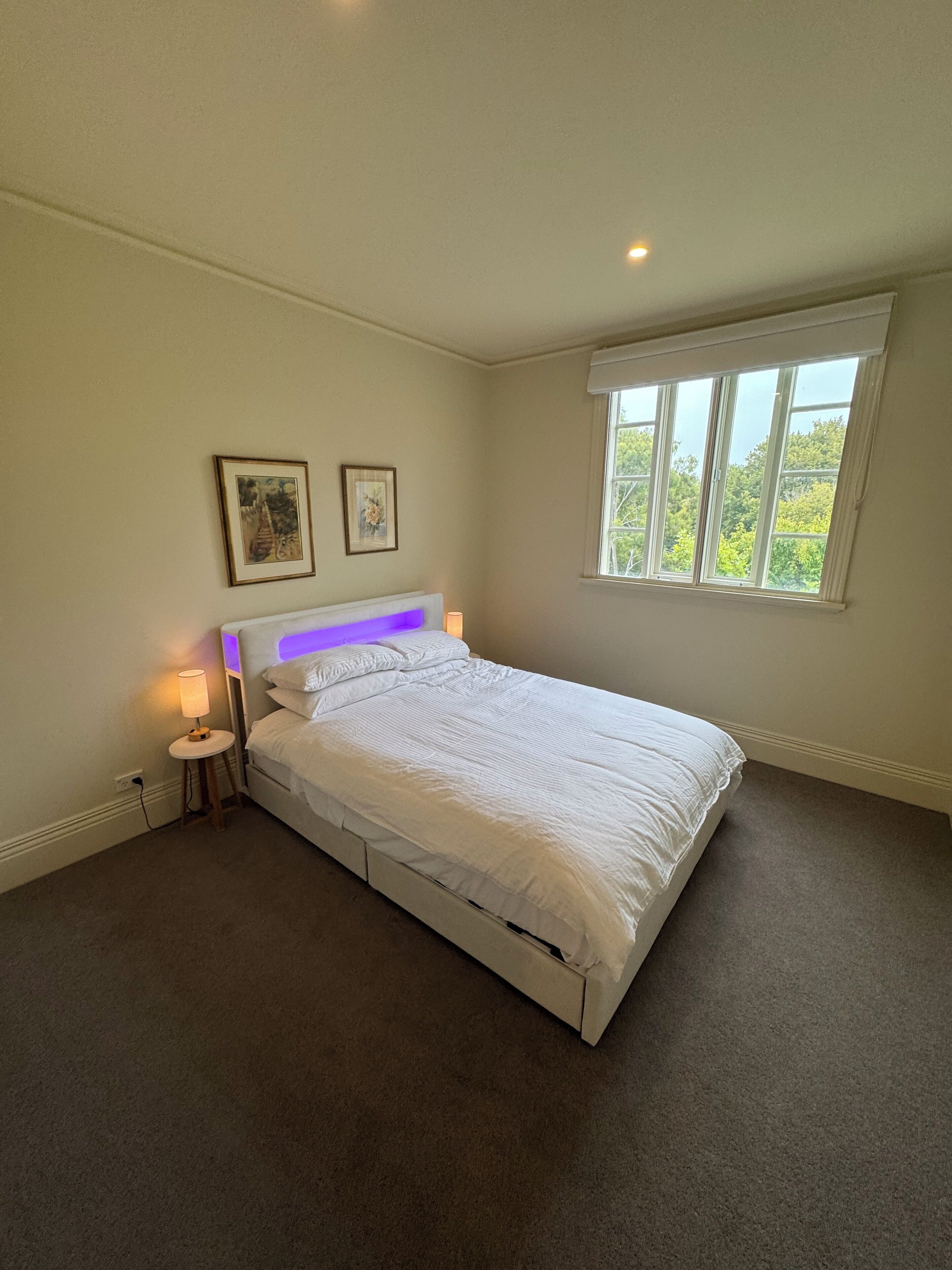 A bedroom with a bed, a nightstand, a lamp, a window, and two framed pictures on the wall.