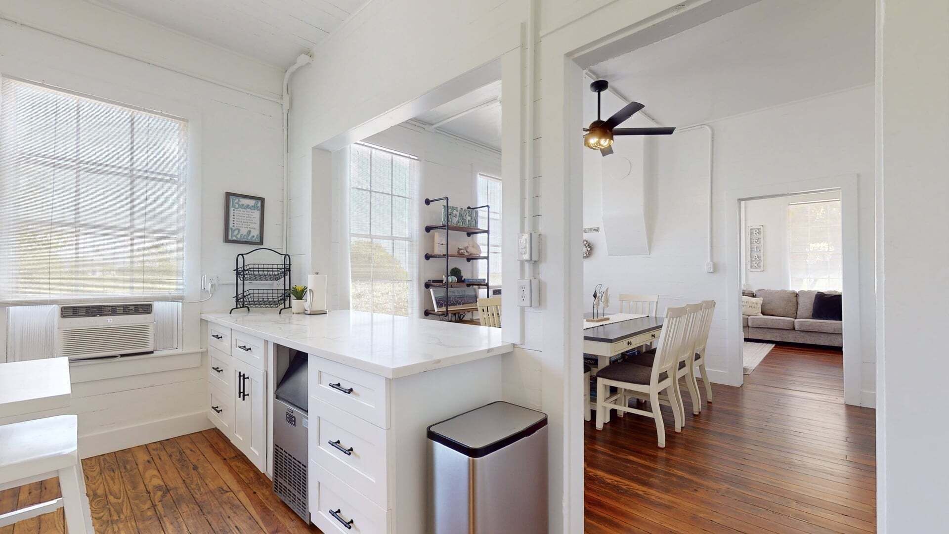A kitchen with white cabinets, a white countertop, a black trash bin, and a dining area with a table and chairs.