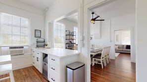 A kitchen with white cabinets, a white countertop, a black trash bin, and a dining area with a table and chairs. - Shiloh By The Sea 5 Bedroom Home (Port Bolivar)