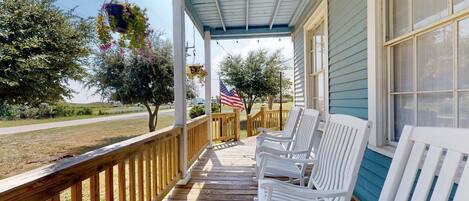 A covered porch with white chairs, a hanging flower pot, and an American flag.