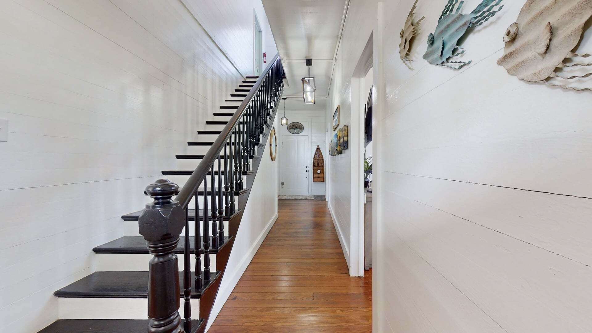 A narrow hallway with a wooden floor, a black staircase with a balustrade, and a white door at the end.