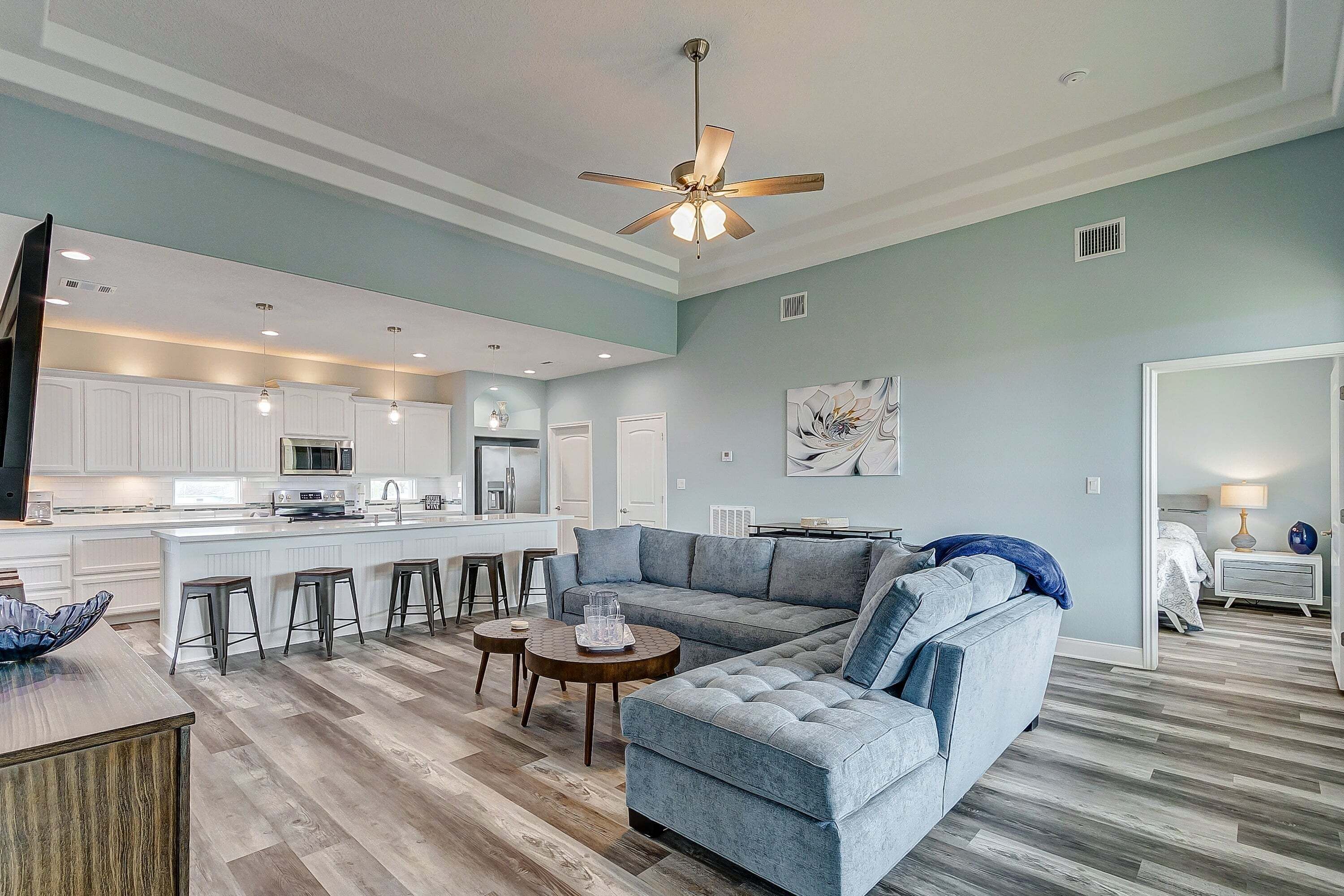 Un salon moderne avec un canapé d’angle, un ventilateur de plafond et un coin cuisine à l’arrière-plan.