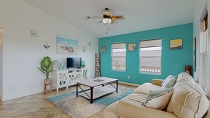 A living room with a teal wall, a ceiling fan, a TV, a coffee table, a sofa, and a window with a view of a wooden deck. - Casa Siwichi 2 Bedroom Home (Port Bolivar)