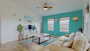 A living room with a teal wall, a ceiling fan, a TV, a coffee table, a sofa, and a window with a view of a wooden deck. - Casa Siwichi 2 Bedroom Home (Port Bolivar)