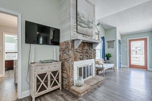 Una sala de estar con chimenea de piedra, televisor montado y puerta que da a otra habitación.