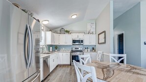 A modern kitchen with white cabinets, stainless steel appliances, and a dining area with a wooden table and white chairs. - The Showboat 3 Bedroom Home (Port Bolivar)