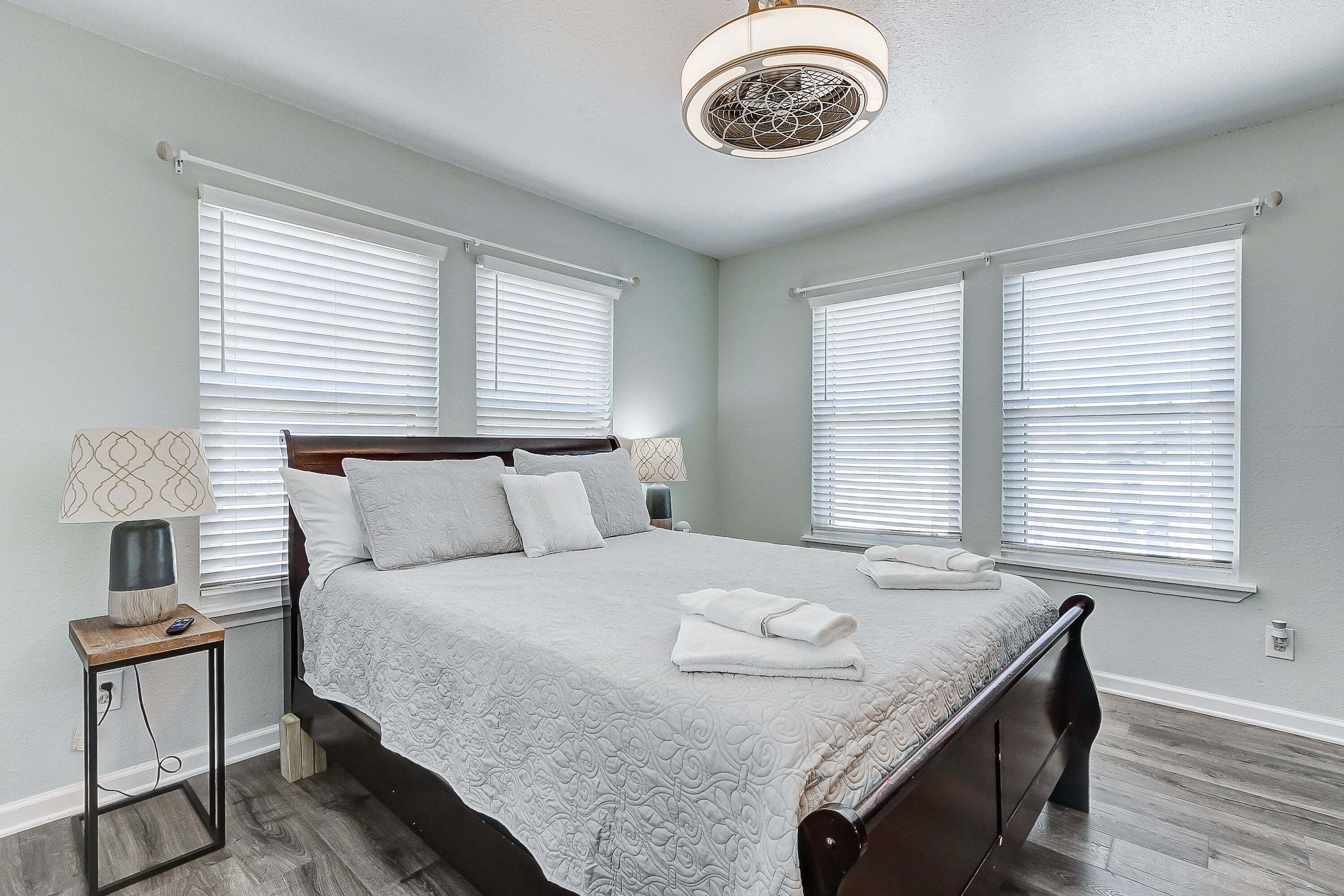 A bedroom with a bed, two bedside tables, a ceiling fan, and large windows with blinds.
