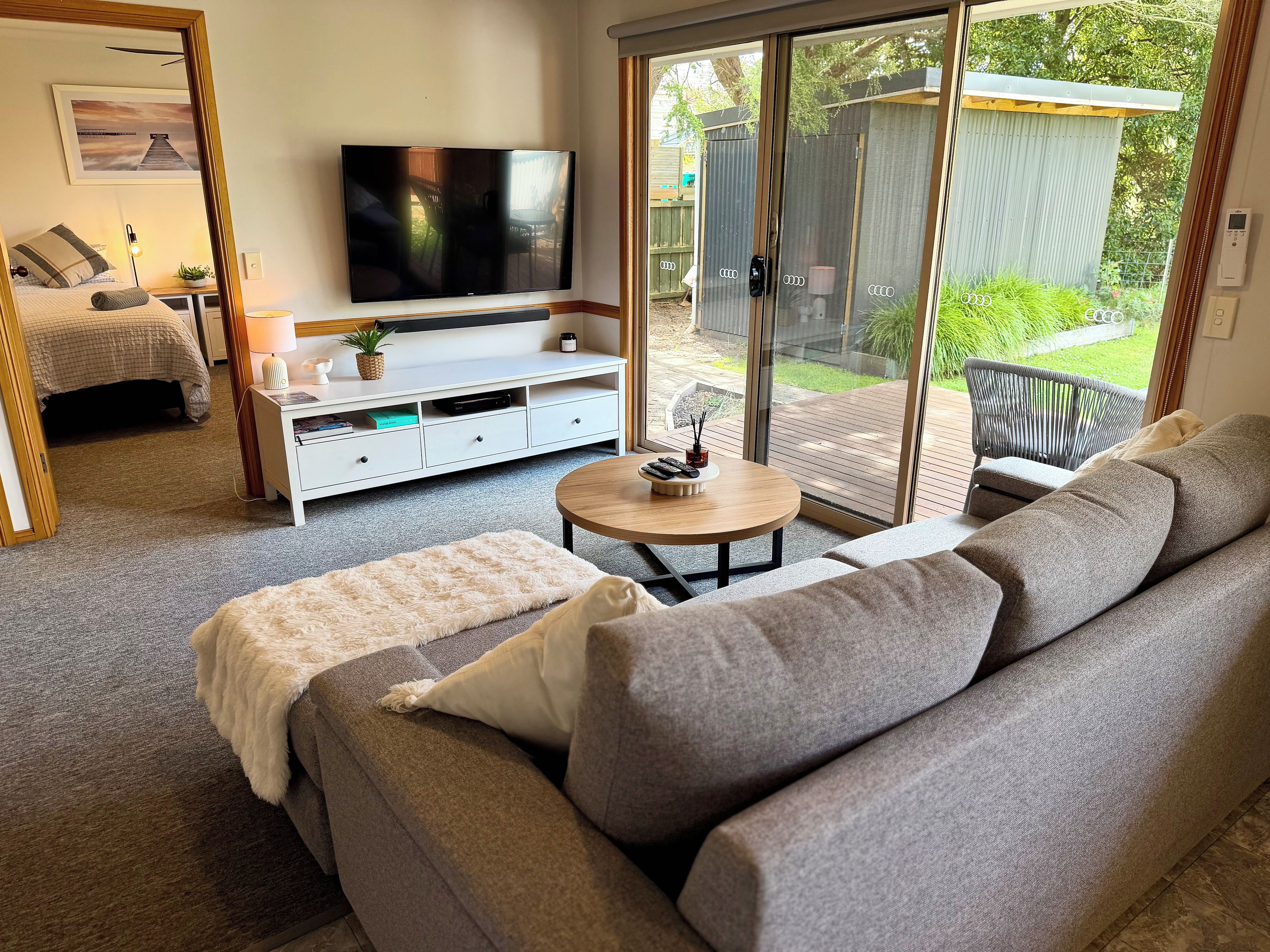 Un salon moderne avec un canapé gris, une table basse, un téléviseur à écran plat et une porte-fenêtre coulissante donnant sur un espace extérieur.