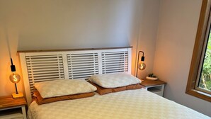A neatly made bed with a quilted white duvet, two pillows, and a folded brown blanket on the bed. There are two bedside tables with lamps, one on each side of the bed.