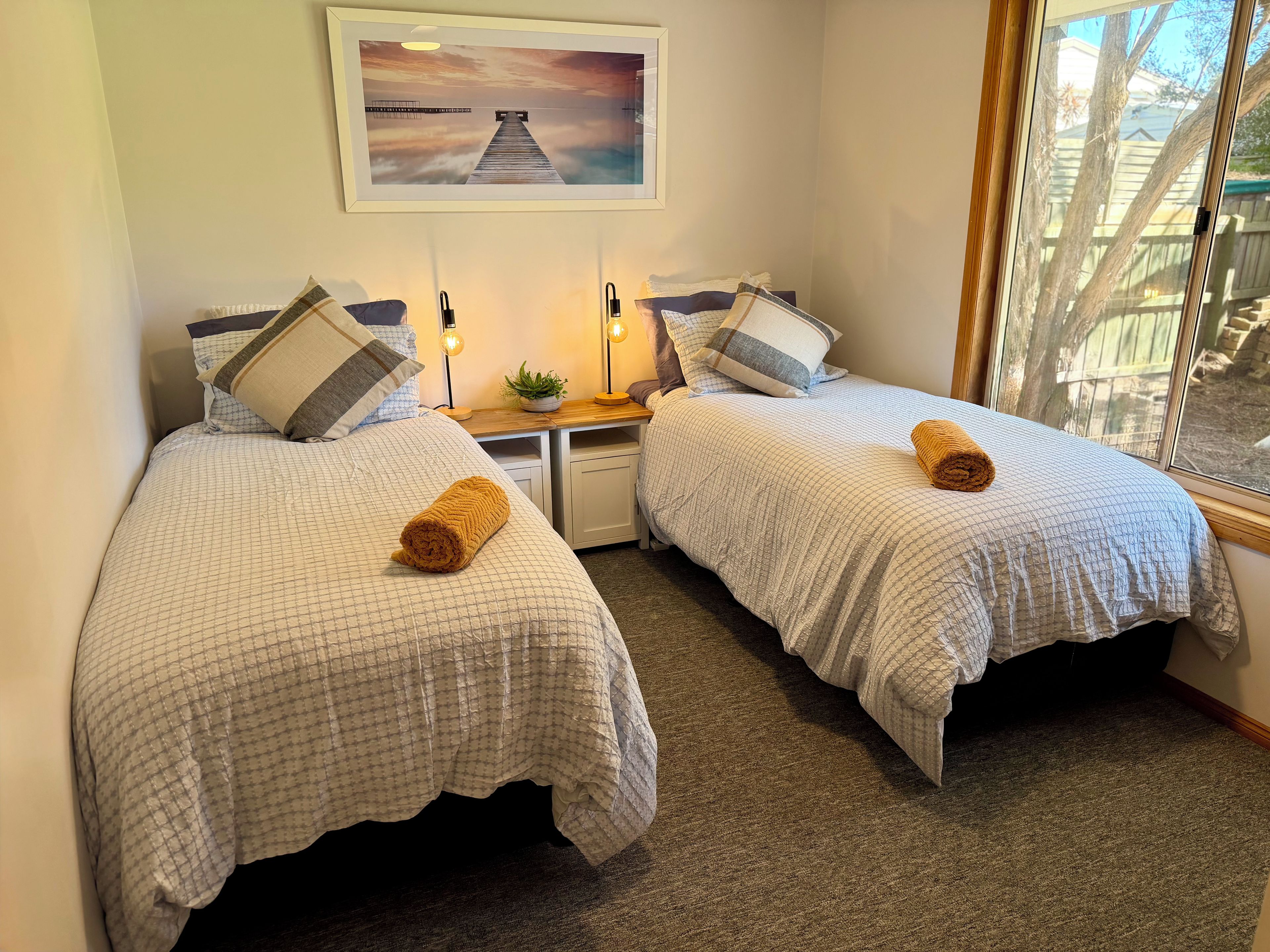 A room with two beds, each with a patterned bedspread and pillows, a small bedside table with a lamp, and a framed picture on the wall.