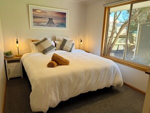 A neatly made bed with pillows and a rolled towel, a bedside table with a lamp, a framed picture on the wall, and a window with a view of trees. - Studio 61 Inverloch - Surfside Retreat (Inverloch)