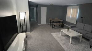 Ein Wohnzimmer mit Flachbildfernseher, einem weißen Sofa, einem Couchtisch und einem Esstisch mit Stühlen.