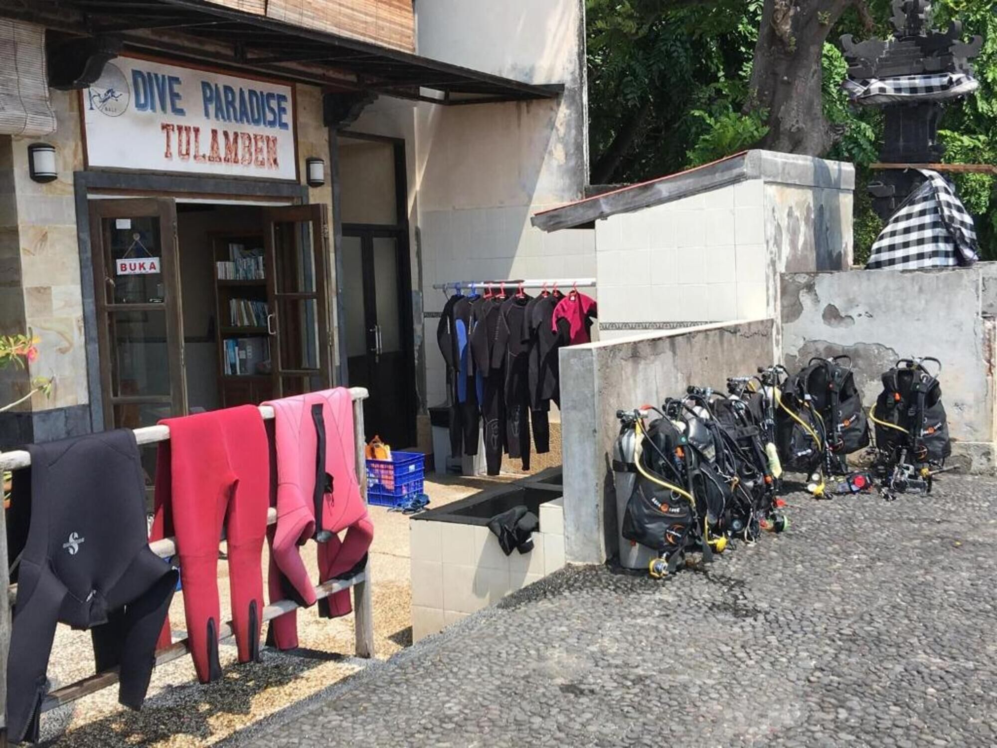 Un magasin de plongée appelé « Dive Paradise Tulamben » expose à l’extérieur son matériel de plongée, notamment des équipements de plongée sous-marine et des combinaisons de plongée.