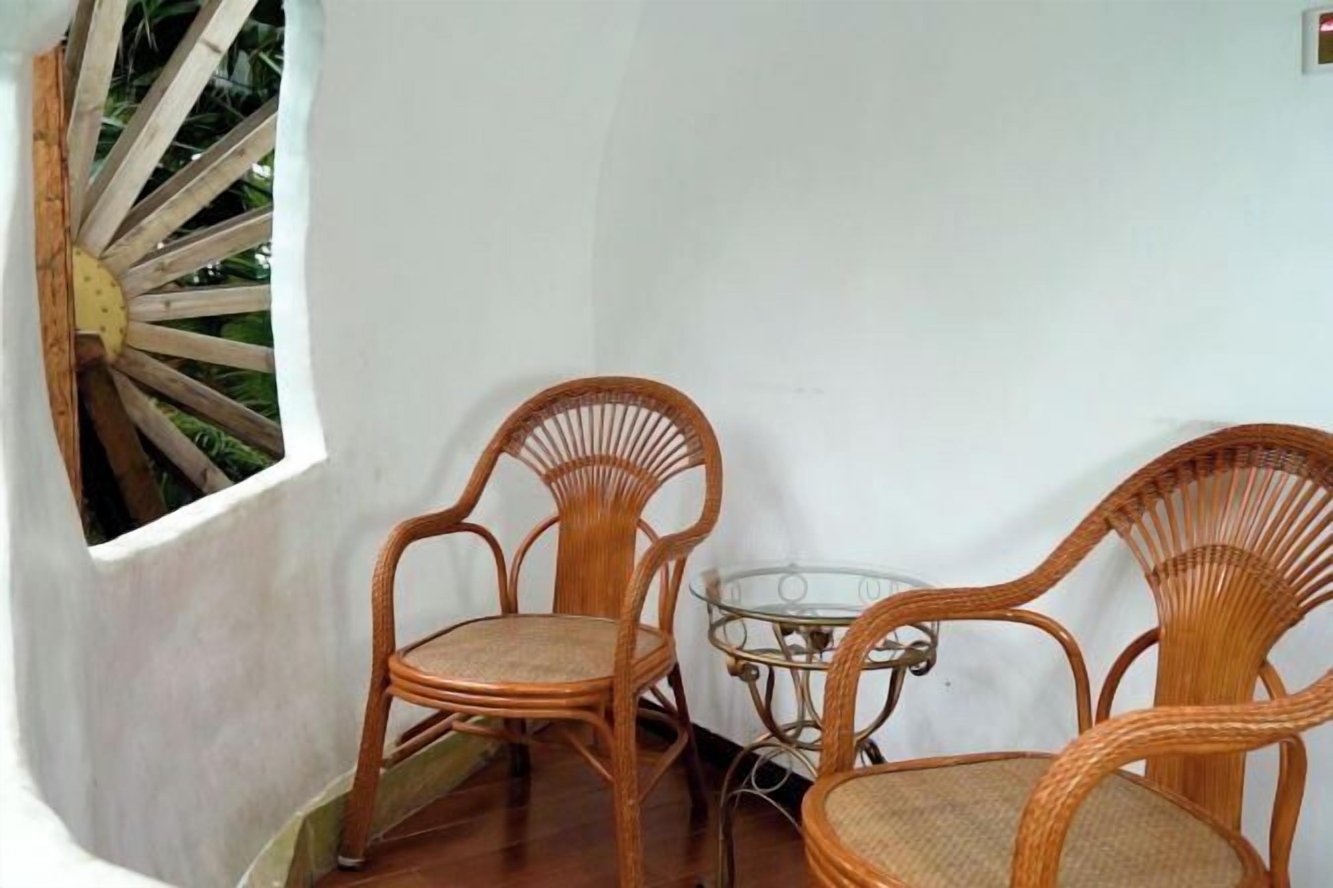 Una habitación con dos sillones de mimbre, una mesa de vidrio y una ventana con marco de madera, junto a una pared blanca.
