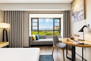 Hotellihuone, jossa on sänky, työpöytä tuolilla, sohva koristetyynyillä, ikkuna, josta avautuu näkymä kaupunkimaisemaan, ja lamppu.
