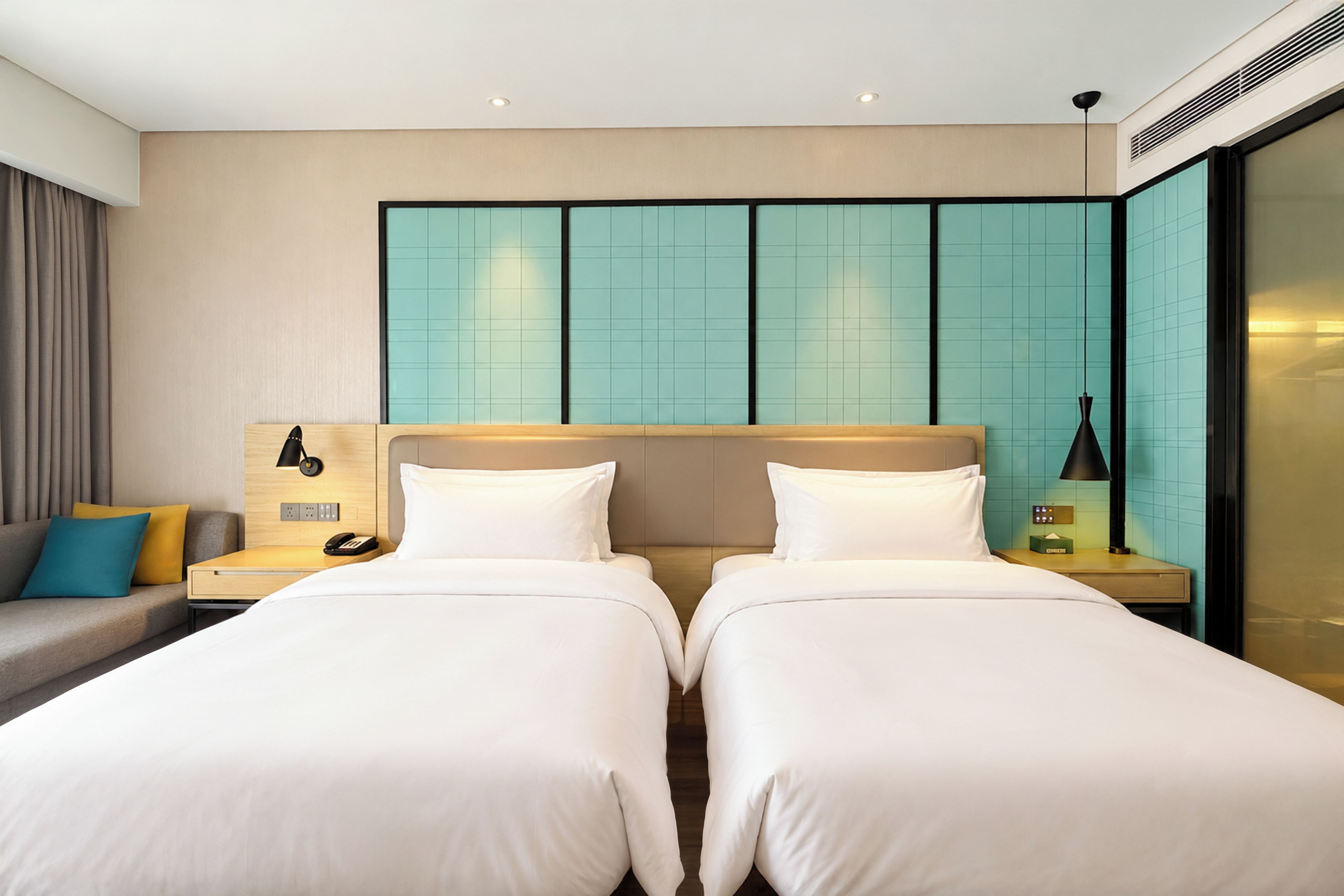 A hotel room with two beds, a sofa, a nightstand with a lamp, and a wall with a grid pattern.